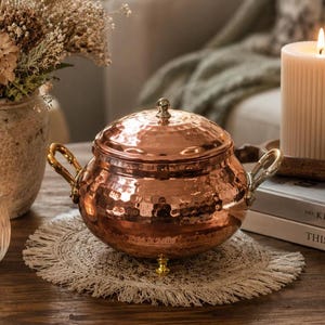 May include: A round, hammered copper pot with a matching lid, featuring two curved brass handles on the sides and three small brass feet at the base. The metallic surface reflects light, showcasing a textured, dimpled finish throughout the exterior.