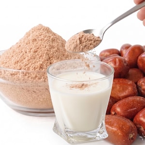 May include: A clear glass bowl filled with brown powder, a glass of white liquid, and a pile of reddish-brown dates. A spoon is pouring brown powder into the glass of white liquid. The image is on a white background.