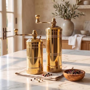 May include: Two cylindrical gold-toned metal spice grinders with manual hand cranks on top. The grinders feature horizontal grooved detailing along the body and a polished reflective finish. A small wooden bowl filled with peppercorns sits nearby.