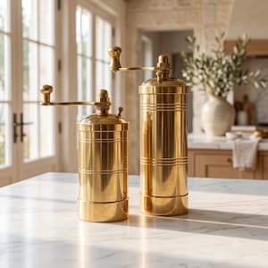 May include: Two cylindrical brass spice grinders of different heights, each featuring a polished metallic finish, a horizontal grooved band detail, and a top-mounted manual crank handle with a small spherical knob for grinding spices.