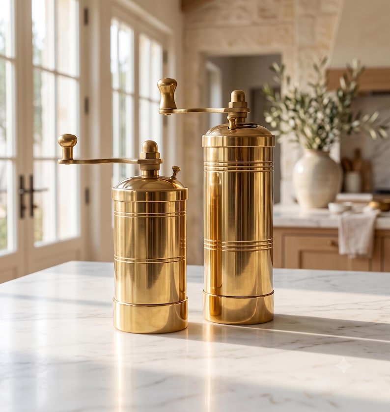 May include: Two cylindrical brass spice grinders of different heights, each featuring a polished metallic finish, a horizontal grooved band detail, and a top-mounted manual crank handle with a small spherical knob for grinding spices.
