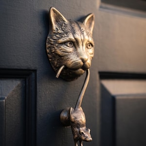 Pode incluir: Um aldravão de porta em metal cor de bronze com a forma de uma cabeça de gato que segura um pequeno rato na boca. A cabeça do gato está montada numa porta escura e o rato serve como a parte oscilante, pendurado pela cauda na boca.