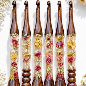 May include: Six crochet hooks arranged vertically, each featuring a dark brown wooden handle at the top and bottom, with a clear resin center section containing embedded dried flowers in shades of pink, yellow, and white.