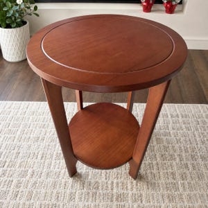 Peut inclure: Une table d'appoint ronde en bois avec une finition brun chaud, dot&eacute;e d'un plateau circulaire lisse avec un d&eacute;tail de bordure l&eacute;g&egrave;rement encastr&eacute; et une &eacute;tag&egrave;re inf&eacute;rieure assortie soutenue par quatre pieds incurv&eacute;s.