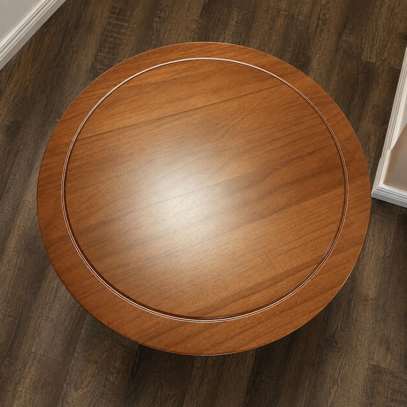 Peut inclure: Une table ronde en bois avec une finition brun moyen lisse. Le plateau pr&eacute;sente un d&eacute;tail d'incrustation circulaire qui cr&eacute;e une bordure subtile autour du centre. Le grain du bois est visible sur toute la surface, montrant des motifs naturels.