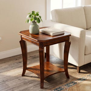 May include: A dark brown wooden side table with a lower shelf and decorative legs. A small green vase with a plant and a book titled "DESIGN The Art of Living" are on the table's surface. The table is next to a beige sofa.