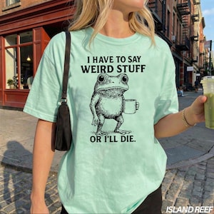 May include: A light mint green short-sleeve t-shirt featuring a black graphic of a frog holding a mug. The text above the frog reads I HAVE TO SAY WEIRD STUFF and the text below the frog reads OR I'LL DIE. The shirt has a crew neckline.