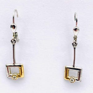 Könnte beinhalten: Ein Paar silber- und goldfarbene Ohrringe. Jeder Ohrring hat einen rechteckigen, goldfarbenen Rahmen mit einem kleinen, runden, klaren Stein am unteren Ende. Die Ohrringe haben einen langen, dünnen Silberstab, der mit einem Silberhaken verbunden ist.