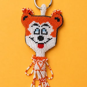 Op de afbeelding: Een kralen sleutelhanger in de vorm van een cartoon vossenkop met oranje, witte en zwarte kralen, voorzien van een zilverkleurige metalen ring. Een metalen liniaal onderaan toont een totale lengte van ongeveer 8,9 cm van boven naar beneden.