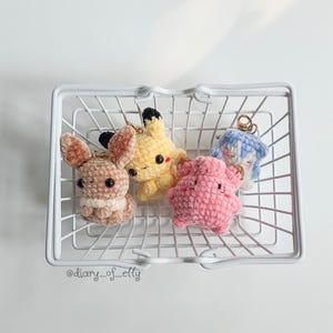 May include: Four small, plush, crocheted animal keychains in a white wire basket. The figures include a brown rabbit, a yellow creature with black-tipped ears, a pink star-shaped character, and a light blue figure. The text @diary_of_elly is visible.