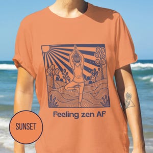 May include: Sunset orange t-shirt with a navy blue graphic of a person in a yoga pose, trees, and sun rays. The text "Feeling zen AF" is below the graphic. A circle with the word "SUNSET" is in the lower left corner.