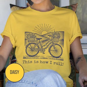 May include: Yellow t-shirt with a navy blue graphic of a bicycle decorated with flowers, baskets, and a rising sun. The text "This is how I roll!" is printed below the bicycle. A yellow circle with the word "DAISY" is in the lower left corner.