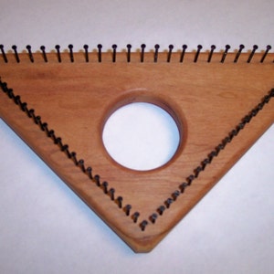 May include: A triangular wooden loom with a circular hole in the centre. The loom has rows of black metal pegs along the edges.