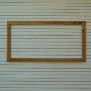 May include: A wooden frame is mounted on a white clapboard wall. The frame is empty and ready for a picture or artwork.