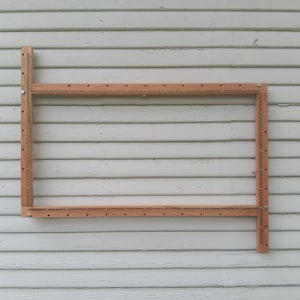 Può includere: Un telaio rettangolare in legno con fori distanziati uniformemente sulla parte superiore e sui lati. Il telaio è realizzato in legno di colore chiaro ed è montato contro un muro di legno dipinto di bianco invecchiato.