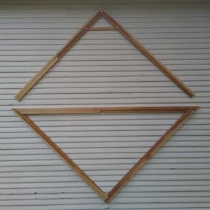 May include: Two wooden triangles, one inside the other, are mounted on a white wooden wall. The triangles are made of thin strips of wood.