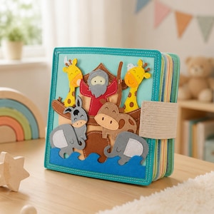 May include: A colorful felt book featuring a scene with Noah's Ark. The cover shows a boat with giraffes, an elephant, a donkey, a hippo, and Noah. The book has a teal border and a beige elastic band closure. The pages have colorful stripes.