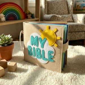 May include: A beige felt book with the words "MY BIBLE" in turquoise and white, with a yellow sun applique. The book has a white handle and a beige strap. The pages are multi-colored.