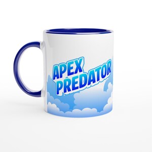 May include: A white ceramic mug with a dark blue handle and interior rim. The exterior features a graphic of light blue clouds and the text APEX PREDATOR in bold, blue, block-style lettering with a white outline, centered on the mug surface.