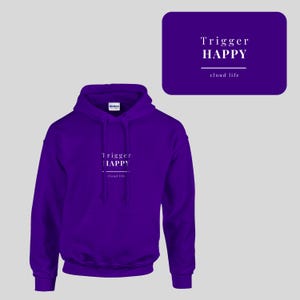 May include: A purple long-sleeve hooded sweatshirt with a front kangaroo pocket and matching drawstrings. The chest features white text that reads Trigger HAPPY with a horizontal line and the words cloud life printed underneath in smaller font.