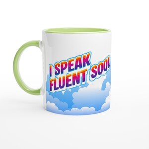 May include: A white ceramic mug with a light green handle and interior rim. The exterior features a graphic of blue clouds at the base and the text I SPEAK FLUENT SOQL in a bold, rainbow-colored gradient font with a white outline and drop shadow.