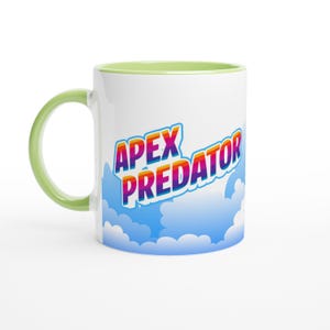 May include: A white ceramic mug with a light green handle and interior rim. The exterior features a graphic of blue clouds and the text APEX PREDATOR in a bold, multi-coloured rainbow gradient font with a white outline and a subtle drop shadow.