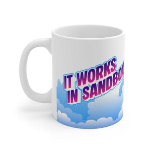 May include: White ceramic mug with a curved handle. The mug features the text "IT WORKS IN SANDBOX" in a gradient of pink and purple, set against a blue and white cloud design. A simple, everyday coffee mug.