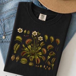 May include: A black t-shirt featuring a botanical illustration of Venus flytrap plants with green leaves, red-tinted traps, and small white flowers. The shirt has a Comfort Colors brand label at the neck, size L, and is displayed with blue denim.