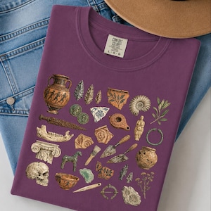 Peut inclure: Un t-shirt couleur prune orné d'une impression graphique représentant divers artefacts archéologiques, dont un vase grec, un crâne, des outils en pierre, des tessons de poterie, des pièces de monnaie, une truelle et un fossile.