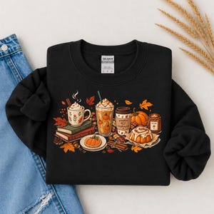 May include: A black crewneck sweatshirt featuring a graphic print of autumn-themed items including mugs of coffee, a pumpkin spice latte, a cinnamon roll, books, pumpkins, and cinnamon sticks, surrounded by scattered orange and red maple leaves.