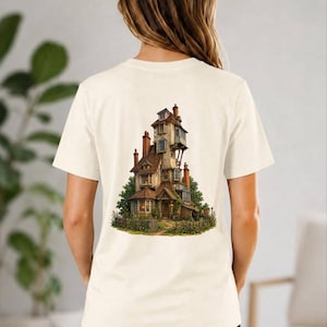 May include: A cream-colored short-sleeve t-shirt featuring a detailed graphic print on the back. The illustration depicts a tall, multi-story, crooked house with brown roofs, multiple chimneys, and surrounding greenery, set within a small fenced garden.