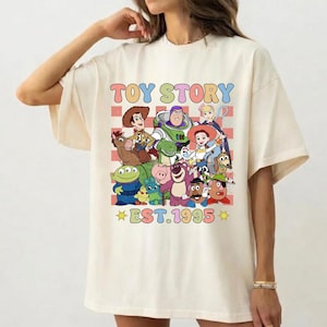 May include: A cream-colored t-shirt featuring a colorful graphic print of Toy Story characters including Woody, Buzz Lightyear, and Jessie. The design includes a red and white checkered background, the text TOY STORY, and EST. 1995.