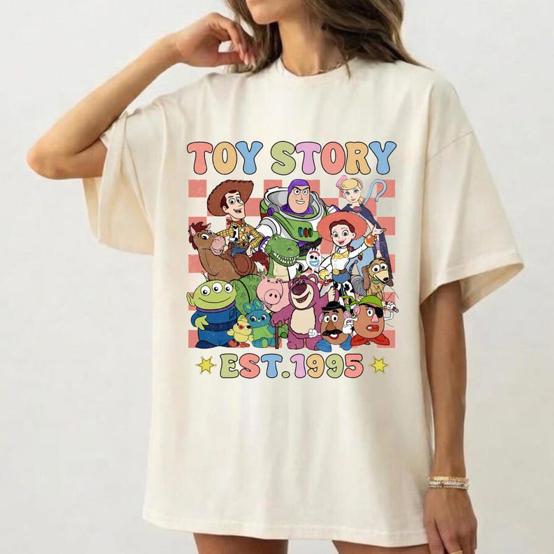 May include: A cream-colored t-shirt featuring a colorful graphic print of Toy Story characters including Woody, Buzz Lightyear, and Jessie. The design includes a red and white checkered background, the text TOY STORY, and EST. 1995.