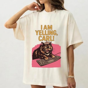 May include: Cream-colored t-shirt featuring a cartoon cat wearing a crown and typing on a keyboard. The text "I AM YELLING, CARL!" is printed above the cat illustration in yellow and brown. The cat is set against a pink background.