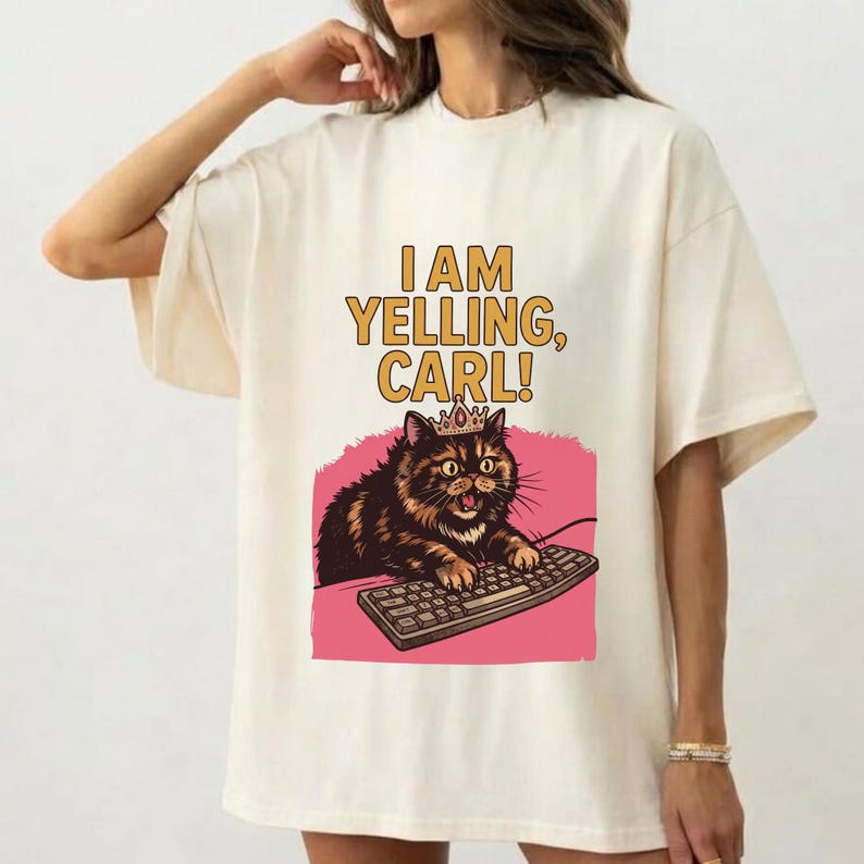 May include: Cream-colored t-shirt featuring a cartoon cat wearing a crown and typing on a keyboard. The text "I AM YELLING, CARL!" is printed above the cat illustration in yellow and brown. The cat is set against a pink background.