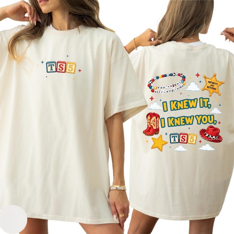 May include: An off-white t-shirt featuring a small TS5 block graphic on the front. The back displays a colorful design with a beaded bracelet, a sheriff star, cowboy boots, a hat, and the text I KNEW IT, I KNEW YOU, TS5, and INFINITY AND BEYOND.