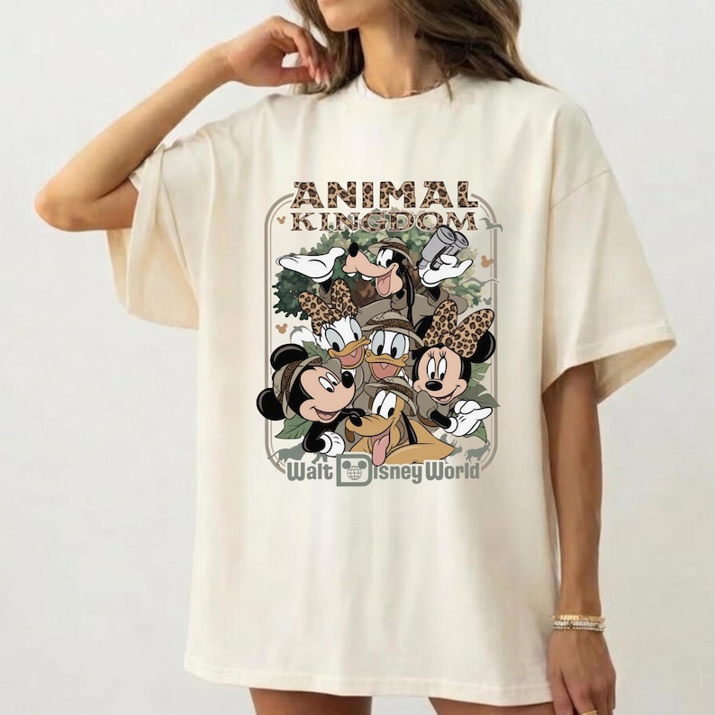 Puede incluir: Camiseta color crema con un gr&aacute;fico de personajes de Disney, incluyendo Mickey y Minnie Mouse, Goofy, Donald y Daisy Duck, y Pluto. Tambi&eacute;n se ve el texto "ANIMAL KINGDOM" y "Walt Disney World".