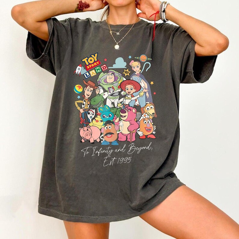 May include: A charcoal gray short-sleeve t-shirt featuring a colorful graphic print of Toy Story characters including Woody, Buzz Lightyear, and Jessie. The text reads Toy Story Land, To Infinity and Beyond, and Est 1995 in white and colored fonts.
