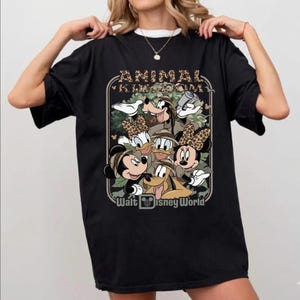 Puede incluir: Camiseta negra con un gr&aacute;fico de personajes de Disney, incluyendo Mickey y Minnie Mouse, Goofy, Donald Duck y Daisy Duck, con el texto "ANIMAL KINGDOM" y "Walt Disney World". Los personajes llevan sombreros de safari.