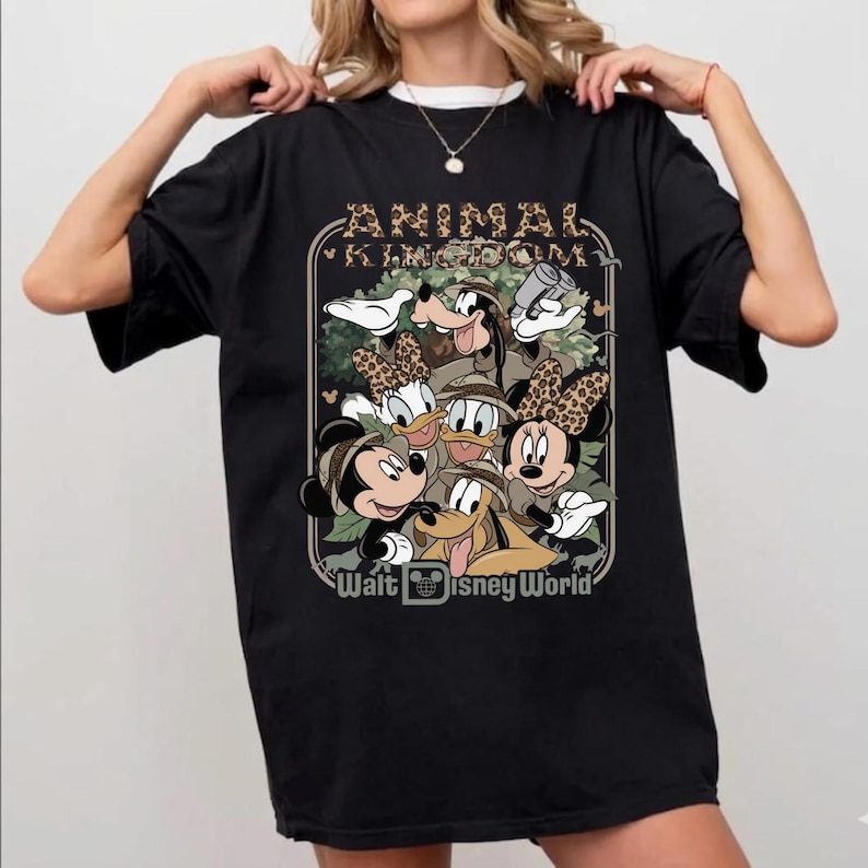Puede incluir: Camiseta negra con un gr&aacute;fico de personajes de Disney, incluyendo Mickey y Minnie Mouse, Goofy, Donald Duck y Daisy Duck, con el texto "ANIMAL KINGDOM" y "Walt Disney World". Los personajes llevan sombreros de safari.