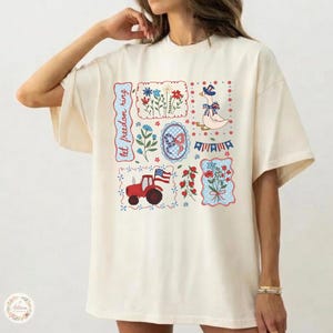 Può includere: T-shirt color crema con un design patchwork con illustrazioni patriottiche. Il design include un trattore rosso, un'oca con un cappello, fiori e la scritta "let freedom ring". Il tema generale è Americana.