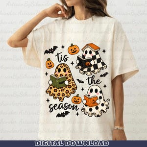 May include: A cream-colored t-shirt featuring a graphic of three ghosts reading books, surrounded by small pumpkins, bats, and stars. The text "tis the season" is printed in black script. The ghosts have leopard print, checkered, and polka dot patterns.