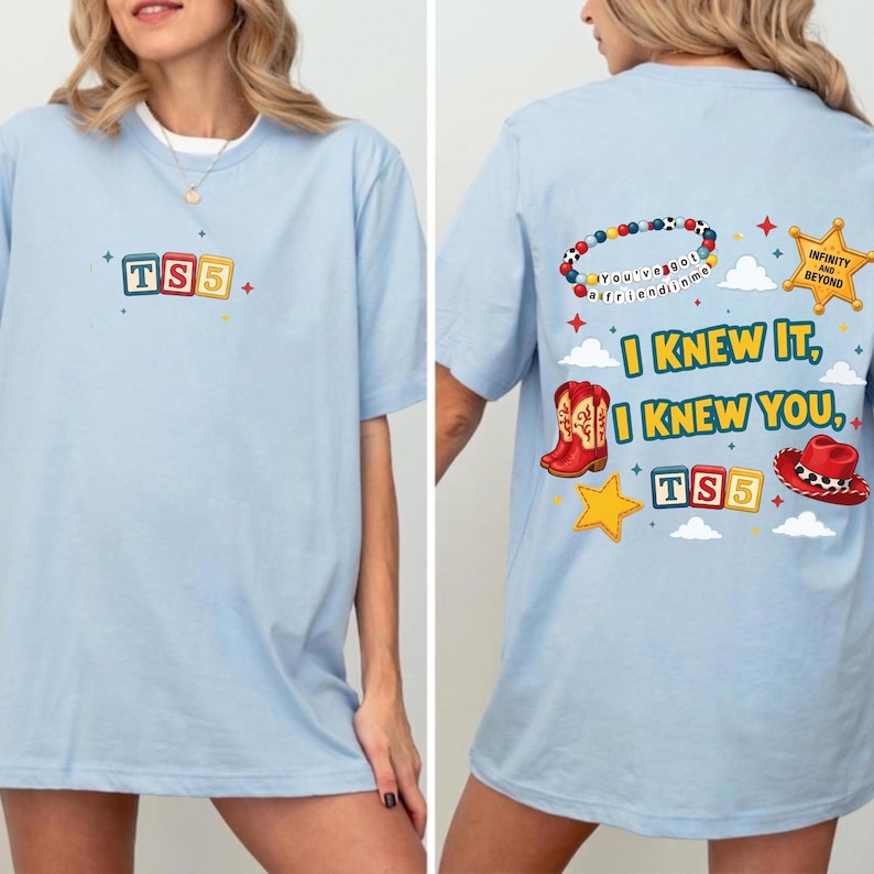 May include: A light blue t-shirt featuring a small graphic on the front with alphabet blocks spelling TS5. The back displays a large graphic with alphabet blocks, a beaded bracelet, a sheriff badge, cowboy boots, and a red cowboy hat.