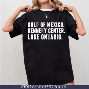 Op de afbeelding: Een zwart T-shirt met korte mouwen en witte tekst die GULF OF MEXICO, KENNEDY CENTER, LAKE ONTARIO leest. De letters F, D en T zijn gemarkeerd in een andere letterstijl binnen de woorden, wat een verticale reeks vormt.