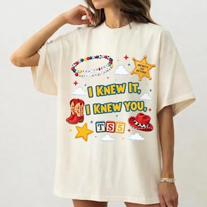 Może przedstawiać: Kremowy t-shirt z nadrukiem graficznym przedstawiającym kolorową bransoletkę z koralik&oacute;w, ż&oacute;łtą gwiazdę szeryfa, czerwone kowbojki, czerwony kapelusz kowbojski oraz klocki z napisem TS5. Tekst głosi I KNEW IT, I KNEW YOU oraz INFINITY AND BEYOND.