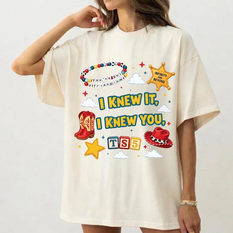 Może przedstawiać: Kremowy t-shirt z nadrukiem graficznym przedstawiającym kolorową bransoletkę z koralik&oacute;w, ż&oacute;łtą gwiazdę szeryfa, czerwone kowbojki, czerwony kapelusz kowbojski oraz klocki z napisem TS5. Tekst głosi I KNEW IT, I KNEW YOU oraz INFINITY AND BEYOND.