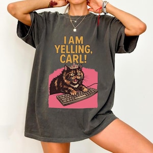 May include: Dark gray t-shirt with the text "I AM YELLING, CARL!" above a cartoon cat wearing a crown and typing on a keyboard. The cat is brown and black with a pink background.