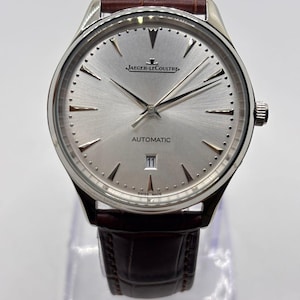 May include: A silver-toned Jaeger-LeCoultre automatic wristwatch with a brown leather band. The watch face is silver with silver hands and markers. The watch has a date display at the 3 o'clock position and the words "AUTOMATIC" and "SWISS MADE" are visible.