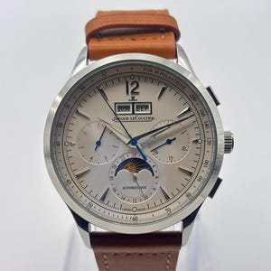 Könnte beinhalten: Eine Jaeger-LeCoultre Automatikuhr mit silbernem Zifferblatt, braunem Lederarmband und Mondphasenanzeige. Das Zifferblatt zeigt den Text JAEGER-LECOULTRE, AUTOMATIQUE, SWISS MADE und FOR 30 PULSATIONS sowie ein Datumsfenster mit 2030 SEP.