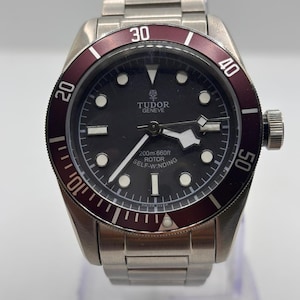 Pode incluir: Um relógio de pulso em aço inoxidável com mostrador preto e moldura bordô. O relógio apresenta marcadores de hora e ponteiros brancos, e o texto "TUDOR GENEVE" é visível no mostrador. A pulseira do relógio também é em aço inoxidável.